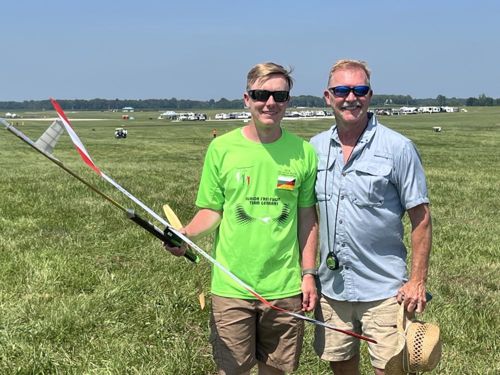 Two people smiling with a model glider on a grassy field.