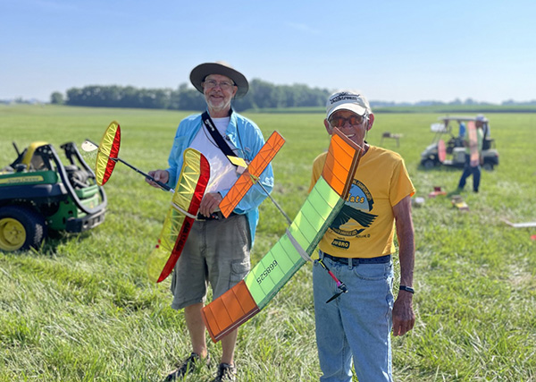 Two men holding colorful model gliders in a grassy field on a sunny day.