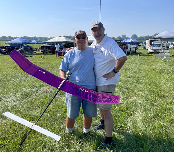 Two people smiling, holding a pink model glider in a grassy field.