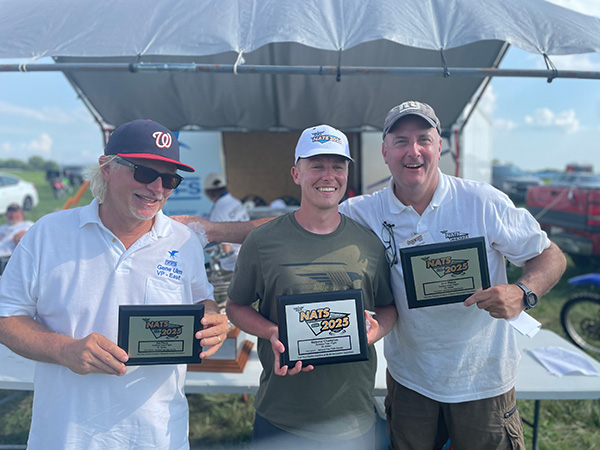 Three men holding awards, smiling under a tent on a sunny day.