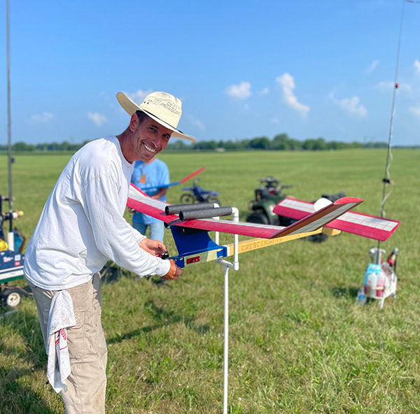 Man in a field smiling, holding a model airplane, under a clear blue sky.