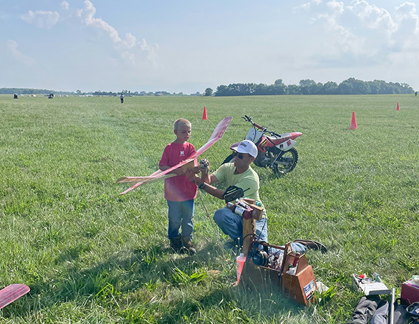 Man and child with model plane on grassy field near small motorcycle.