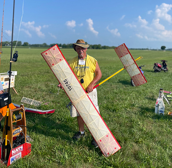 Man in a field holding a large model airplane, wearing a yellow shirt and hat.