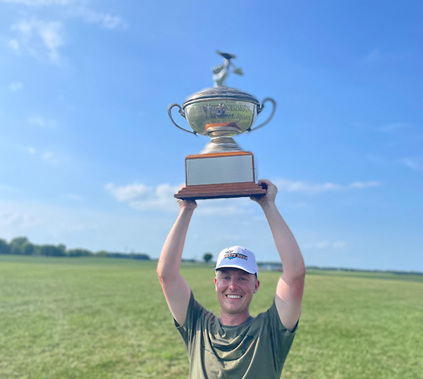 Man smiling and holding a trophy above his head in a grassy field.