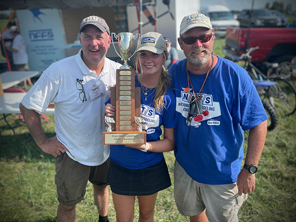 Three people smiling, holding a large trophy outdoors.