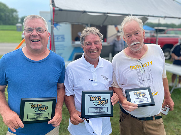 Three men smiling outdoors, holding award plaques, standing side by side.