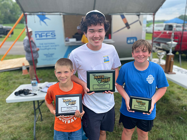 Three boys outdoors holding plaques, smiling at an award event.