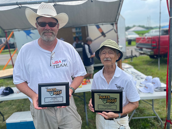 Two men in hats holding awards outdoors with tents in the background.
