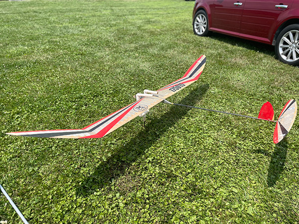 Model glider with red accents on grass near red car.