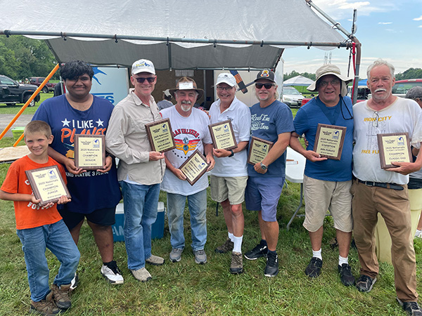Seven people standing outdoors, holding award plaques, smiling in casual attire.