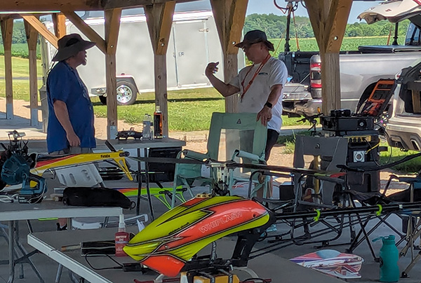 Two men talking under a shelter with model helicopters on tables.
