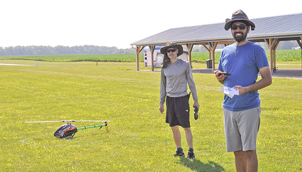 Two people stand on grass with a small helicopter, smiling, under a sunny sky.