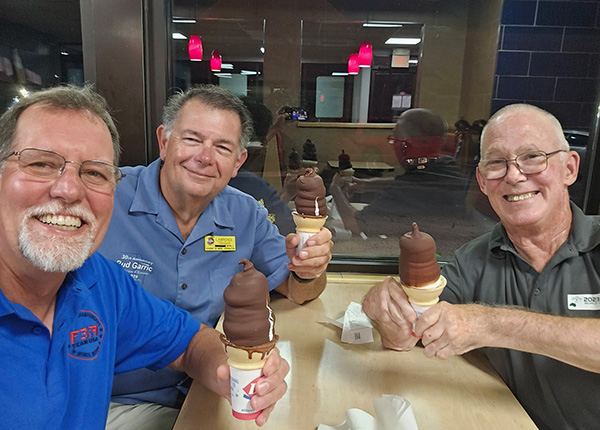 Three smiling men holding chocolate-dipped ice cream cones indoors.