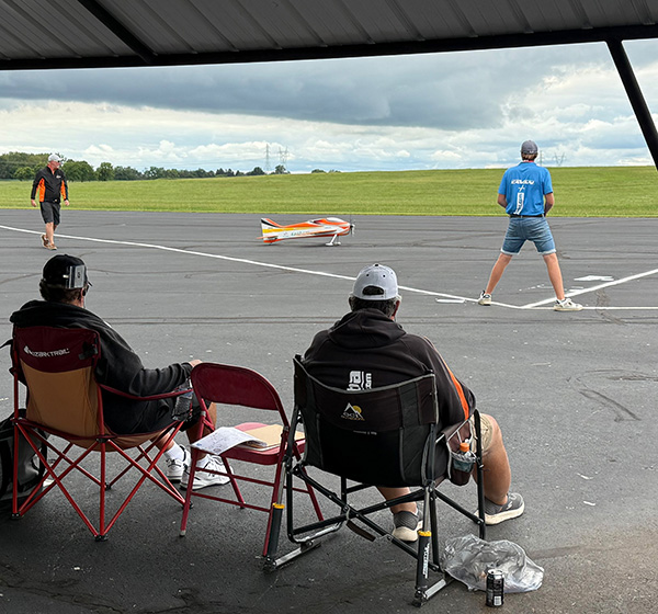 Outdoor model airplane event; people watch a plane on asphalt with grassy field beyond.