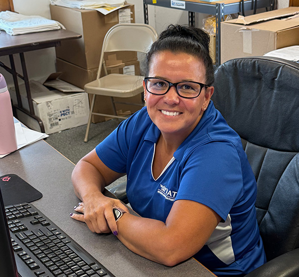 Smiling person in a blue polo shirt seated at a desk with a computer.
