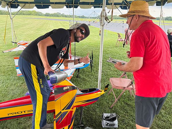 Two people working on model airplanes under a canopy on grass.