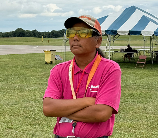 Man in pink shirt with crossed arms stands on grass near a striped tent.