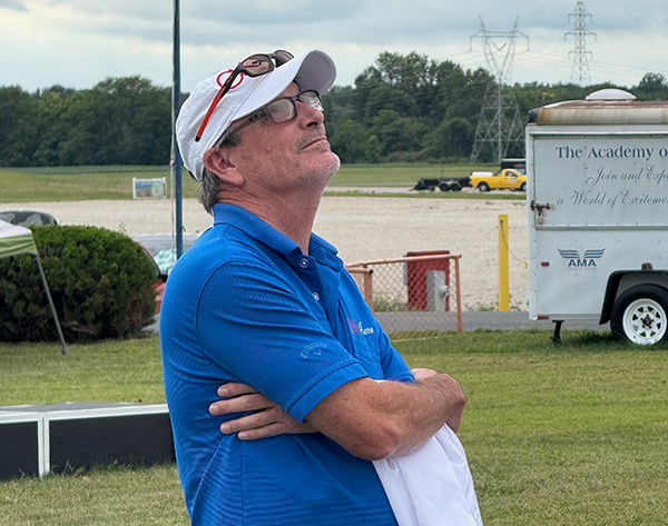 Man in blue shirt and white cap, gazing up with arms crossed outdoors.