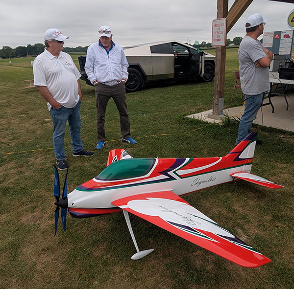 Remote-controlled plane on grass, three men nearby, car in background.