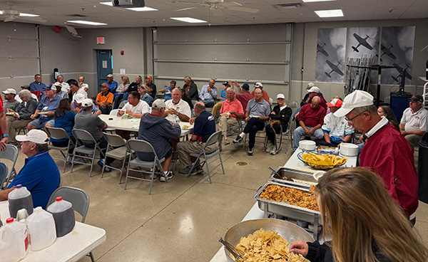 Large group of people seated in a hall, food laid out on tables, casual setting.