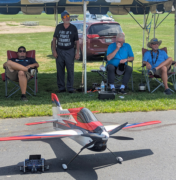Model airplane on ground, four men seated under canopy in background.
