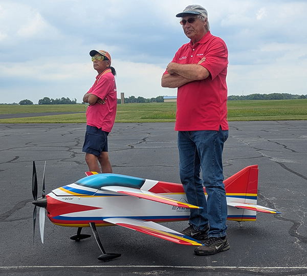 Two men in red shirts stand beside a large model airplane on a runway.