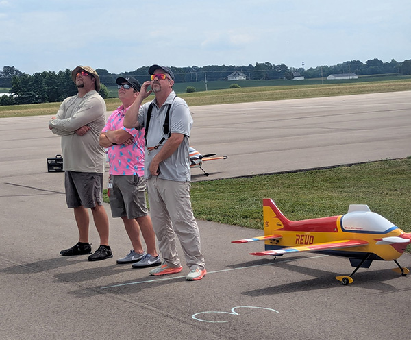 Three men standing on an airstrip, watching a model plane.