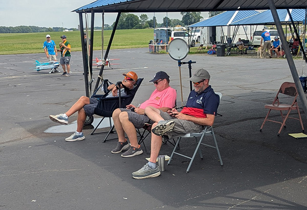 Three people sitting under a canopy on an airfield, watching planes.