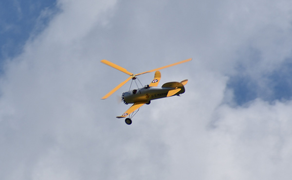 Yellow autogyro flying against a cloudy sky.