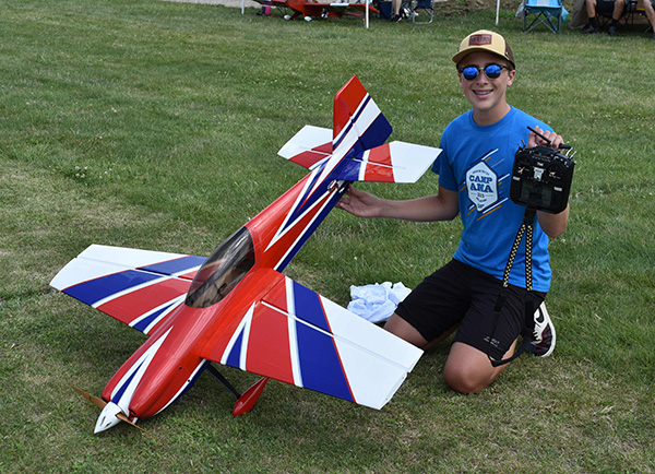 Teen posing with red and blue model airplane on grass, wearing sunglasses and a cap.
