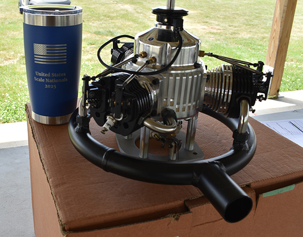 Metal engine model on a box beside a blue tumbler, outdoors.