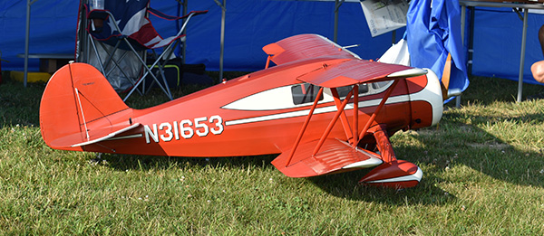 Red model airplane on grass with white stripe and markings, tent and chairs in the background.