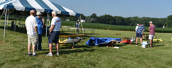 Tent and people gathered on grassy field with model airplanes.
