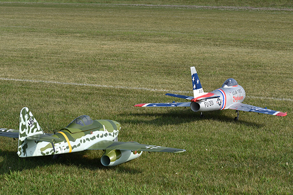 Two model aircraft with colorful markings on grass, one camouflaged, the other silver and blue.