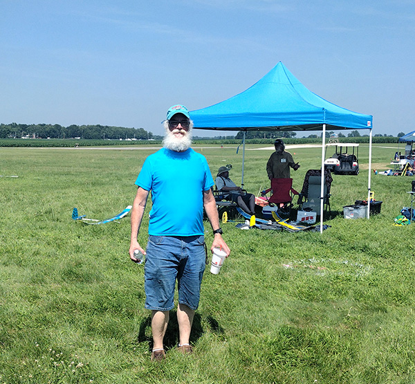 Bearded man in blue outdoors near a blue canopy on a grassy field.
