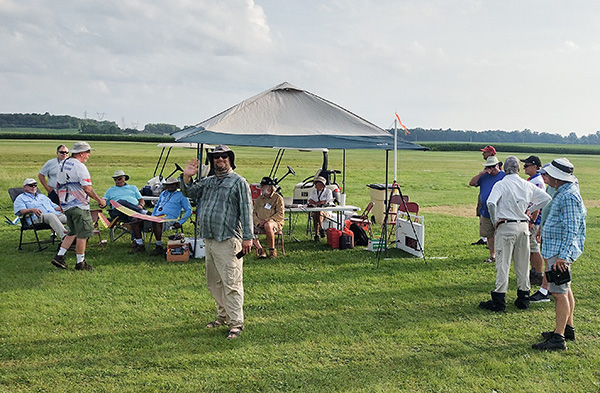 Gathering of people under a canopy at a grassy field, some seated, others standing, with model aircraft.