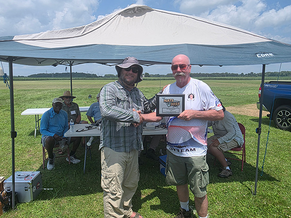 Two men under a canopy holding a framed certificate on a sunny day.