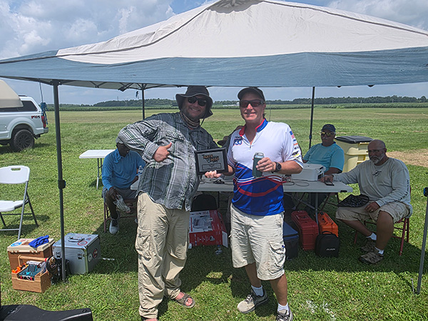 Two men smiling under a canopy, with others seated at a table behind them.