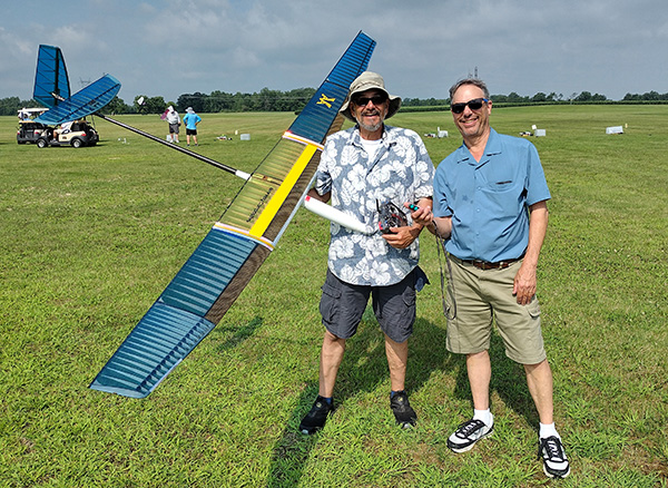 Two smiling men holding a model plane on a grassy field.