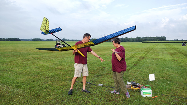 Two people outdoors, one holding a large model aircraft, on a grassy field.