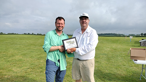Two men smiling, holding a certificate, standing on a grassy field under a cloudy sky.