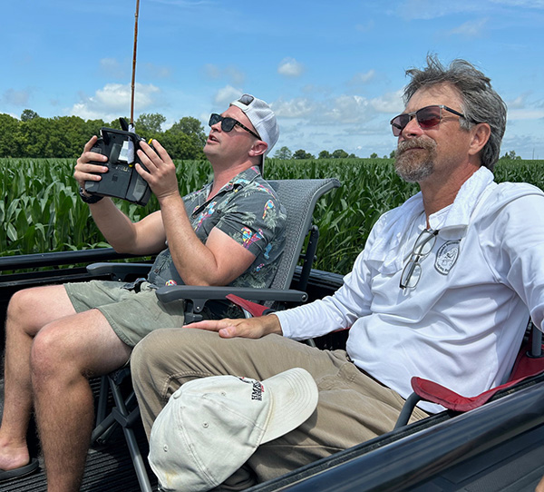 Two men in a field, one standing, operating a device, both appear focused.
