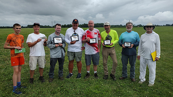 Eight men stand in a grassy field, some holding plaques, under a cloudy sky.