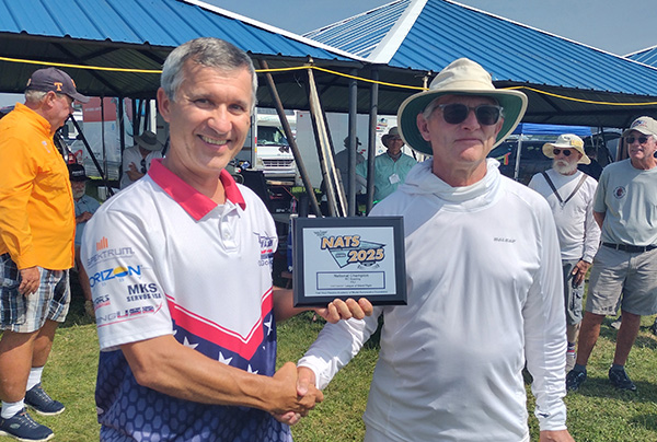 Two men smiling, shaking hands; one holds a framed certificate outdoors.