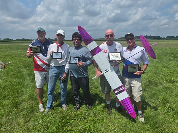 Five people hold plaques and a model plane in a grassy field under a cloudy sky.