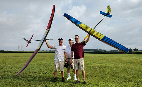 Three men holding large colorful model gliders in an open grassy field.