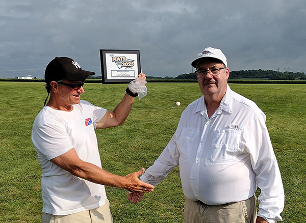 Man holds plaque, smiles with another man on a grassy field, both wearing caps.