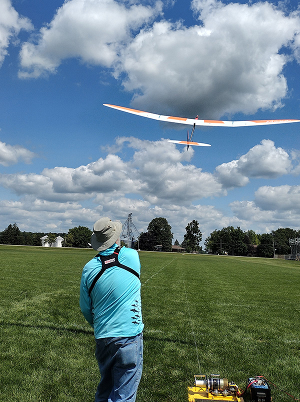 Man launching a model glider in a grassy field under a blue sky with clouds.