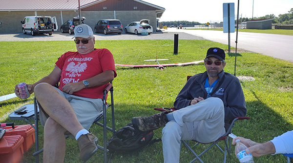 Two men sitting on chairs in a grassy area, relaxed, with parked cars in the background.