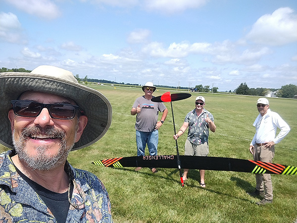 Four men in a field with a large model aircraft; one taking a selfie.